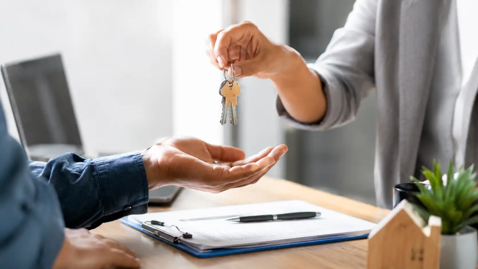 Agente entregando llaves de casa a comprador sobre escritorio con contrato firmado y miniatura de vivienda