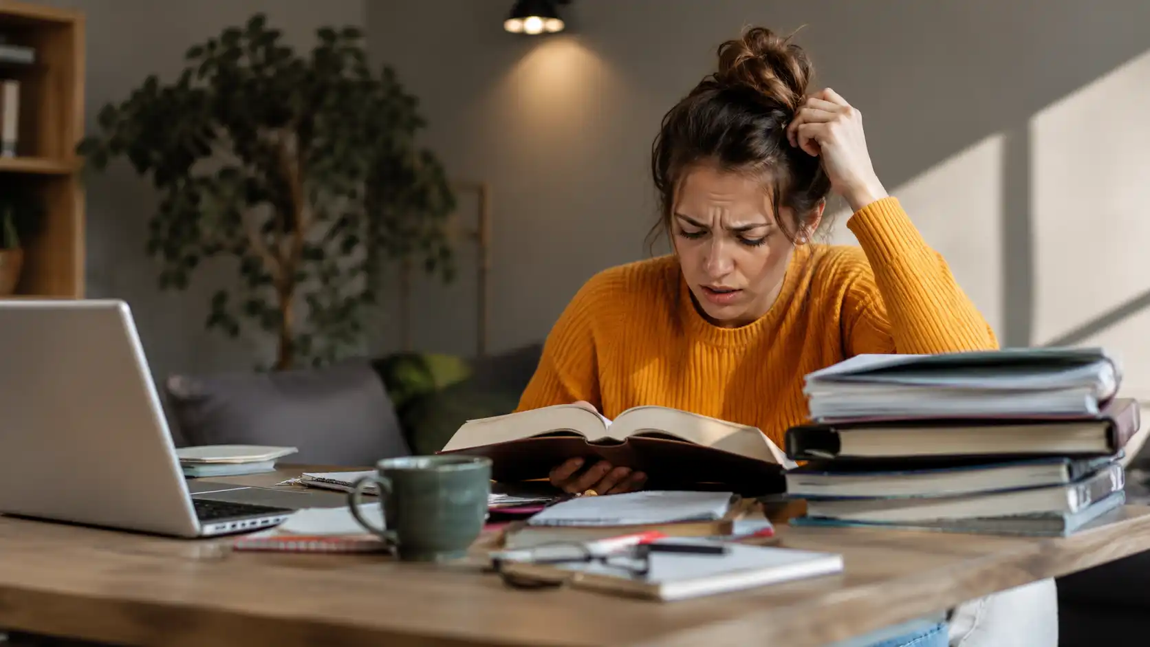 Mujer con suéter naranja confundida leyendo libro rodeada de pila de libros laptop y taza de café en casa